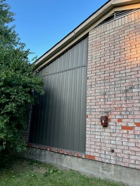 Metal siding on a brick house with a green tree and clear blue sky in the background.
