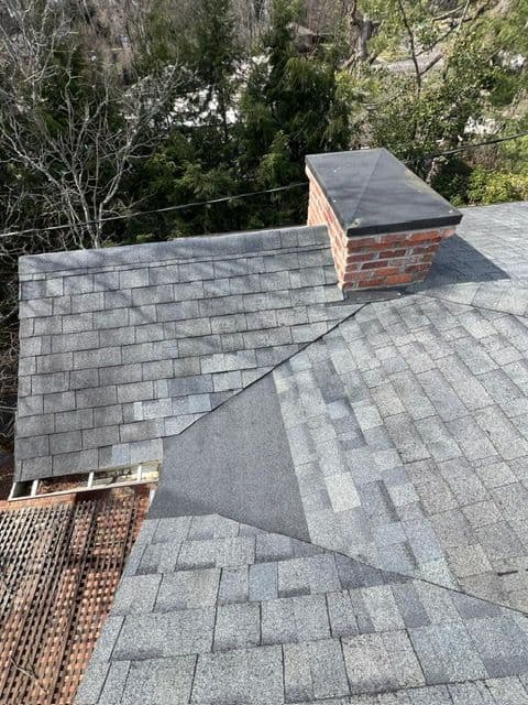 Aerial view of two roof sections with gray shingles and a brick chimney surrounded by trees.