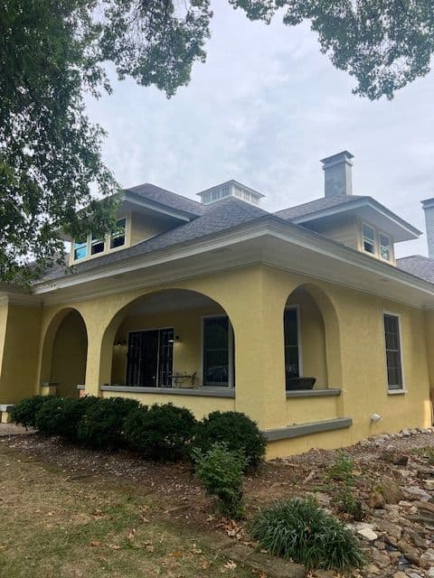Yellow two-story house with arched porch, stone landscaping, and gray roof against cloudy sky.
