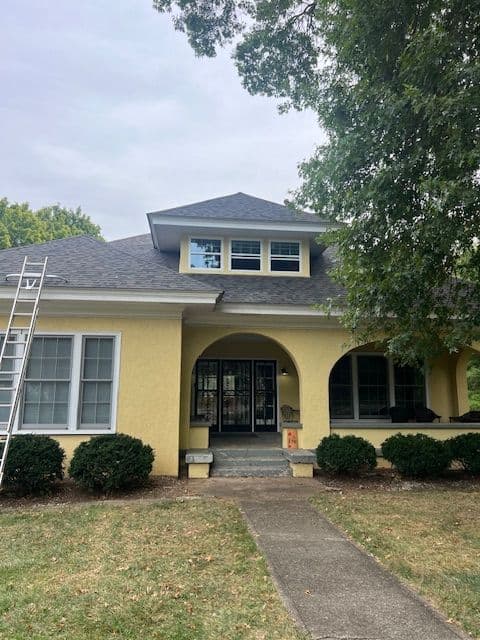 Yellow two-story house with a porch, manicured bushes, and a ladder leaning against the side.