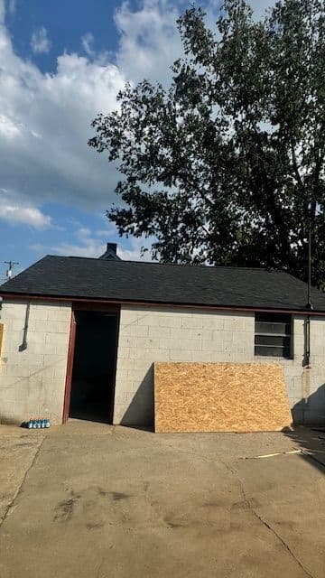 Exterior view of a gray cinder block garage with an open door and plywood in front.