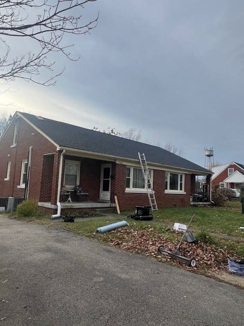 Brick house with new roof installation, ladder propped against the side, and autumn leaves on the lawn.