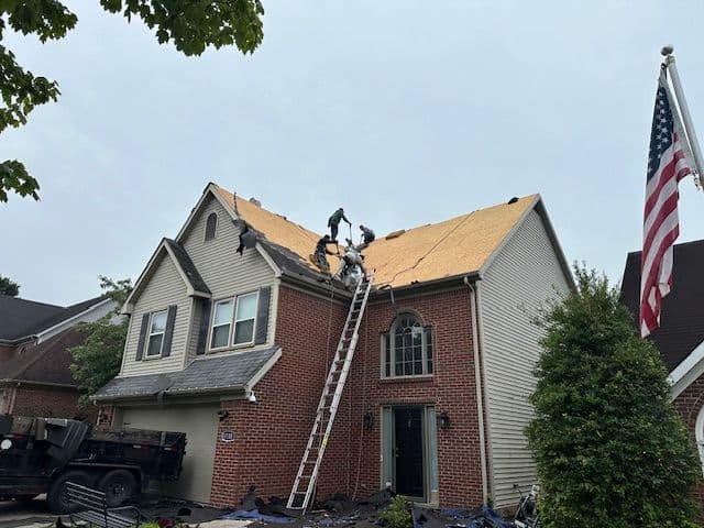Roofing workers installing shingles on a house with an American flag in the foreground.