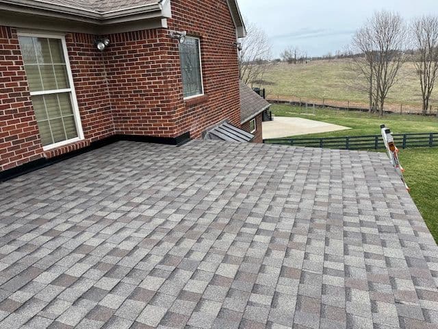 Flat rooftop with gray shingles on a brick house, surrounded by green grass and trees.