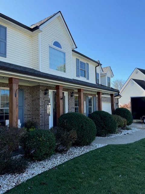 Two-story suburban house featuring a mix of brick and siding, well-manicured shrubs, and a clear sky.