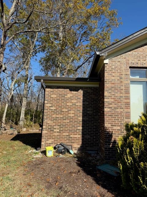 Brick house corner with overhanging roof, surrounded by trees and landscaped yard.