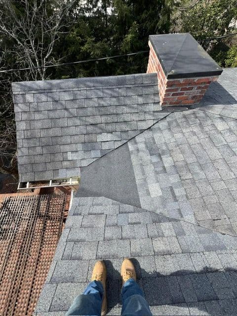 Person standing on a gray shingled roof near a brick chimney and tree line.