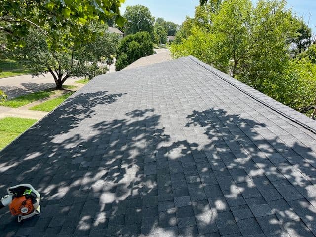 Roof showing gray shingles with sunlight creating shadow patterns, surrounded by trees.