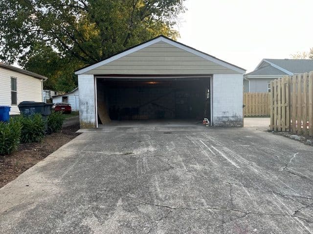 Empty garage with open door and concrete driveway, surrounded by greenery and wooden fence.