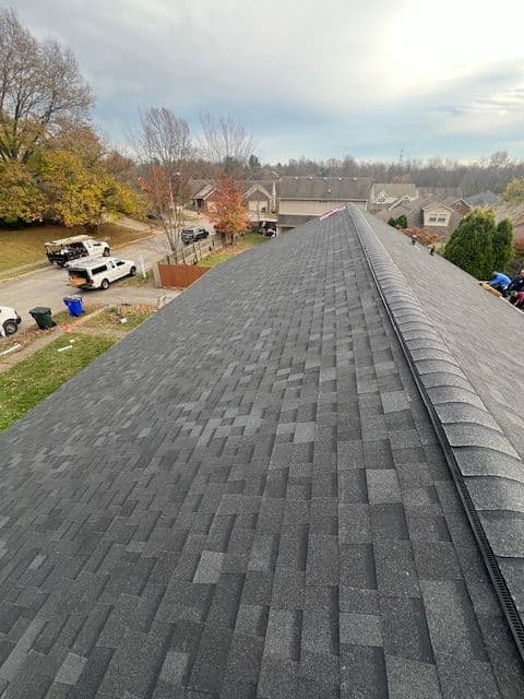 Aerial view of a residential roof, showcasing dark shingles and nearby houses and trees.