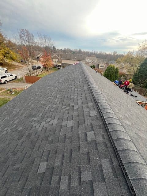Man installing shingles on a sloped gray roof with houses in the background.