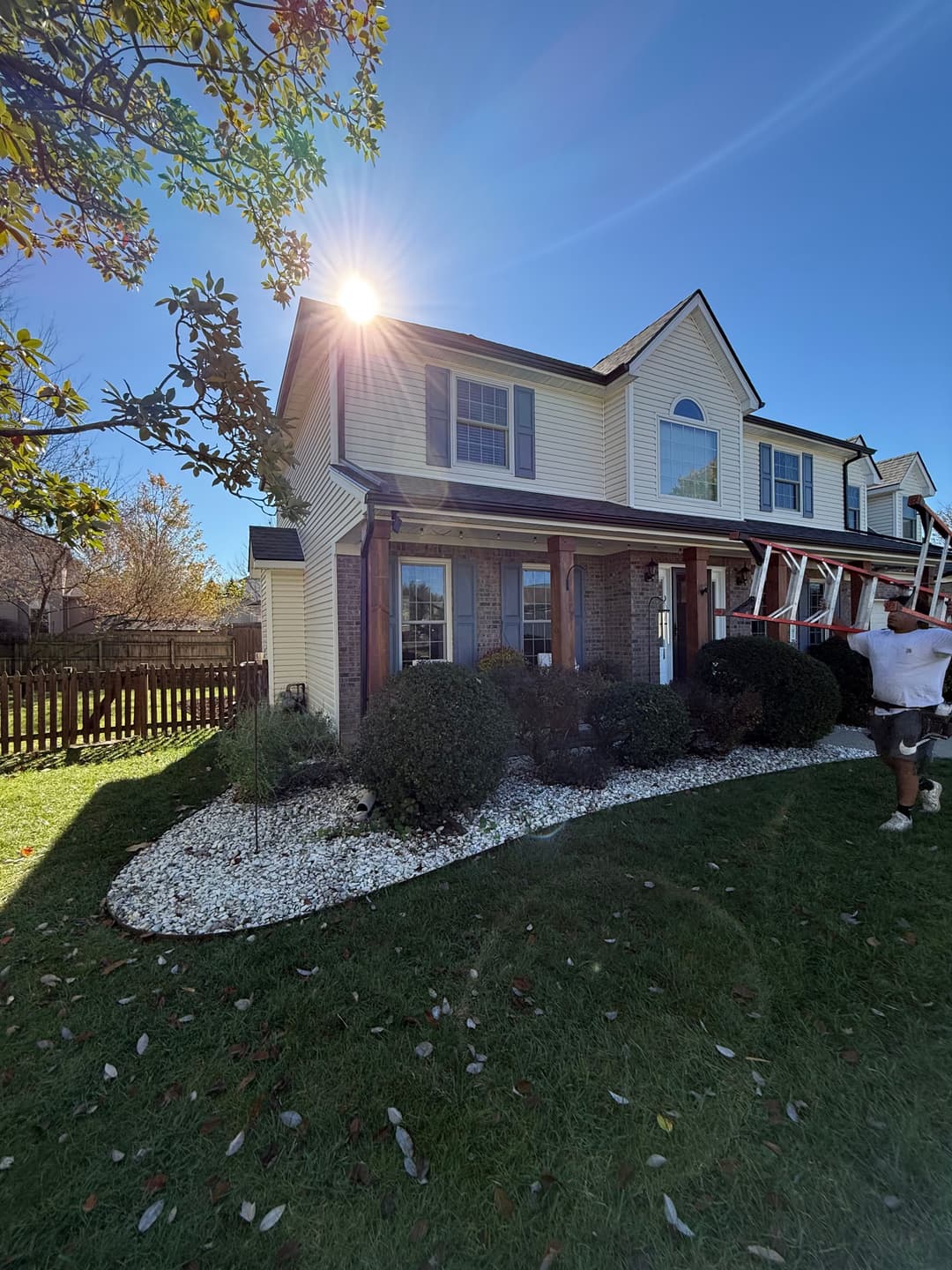 Two-story house with sunlit exterior, landscaped yard, and ladders near the entrance.