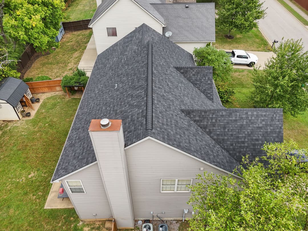 Aerial view of residential house with a dark shingle roof and surrounding greenery.
