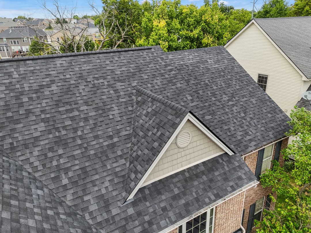 Aerial view of a modern house with gray shingle roof and vibrant green trees nearby.