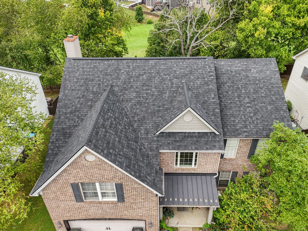 Aerial view of a brick house with a black shingle roof surrounded by greenery.