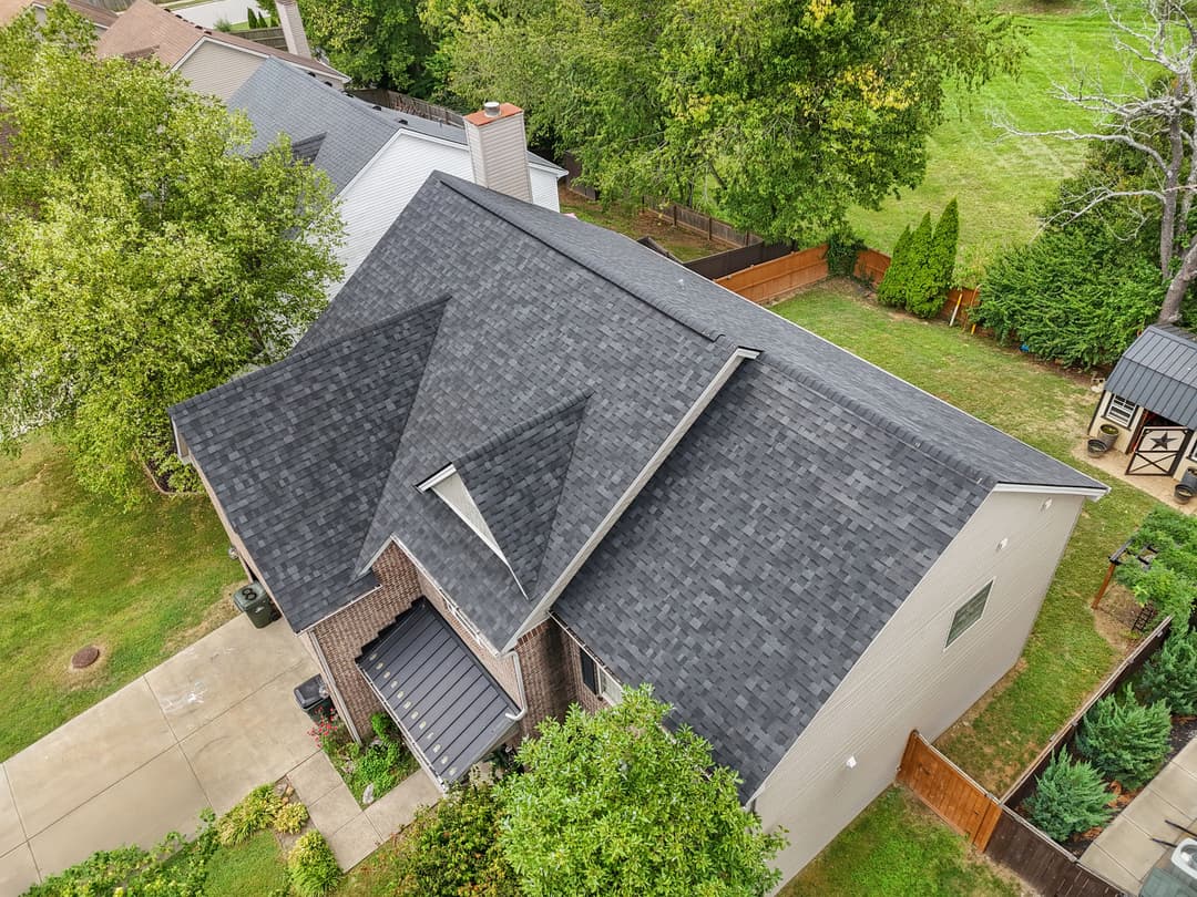 Aerial view of a house with a dark shingle roof surrounded by greenery and a driveway.
