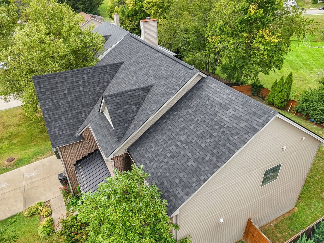 Aerial view of a modern home with a dark shingle roof and lush green surroundings.