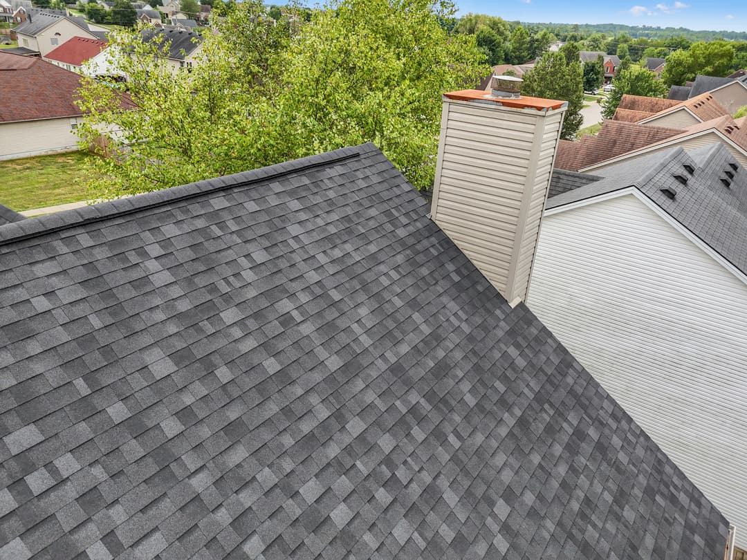 Aerial view of a gray shingle roof with a chimney and surrounding residential homes.