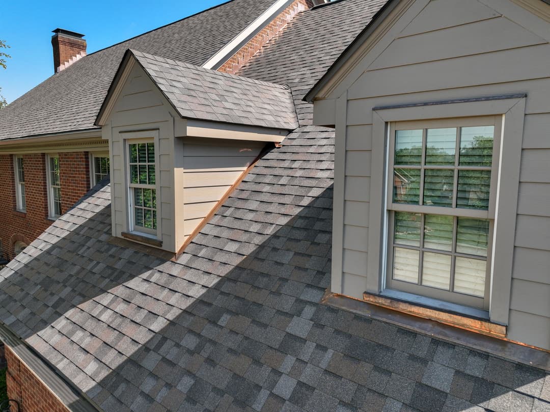 Roofing details of a house with shingles and dormer windows against a clear blue sky.