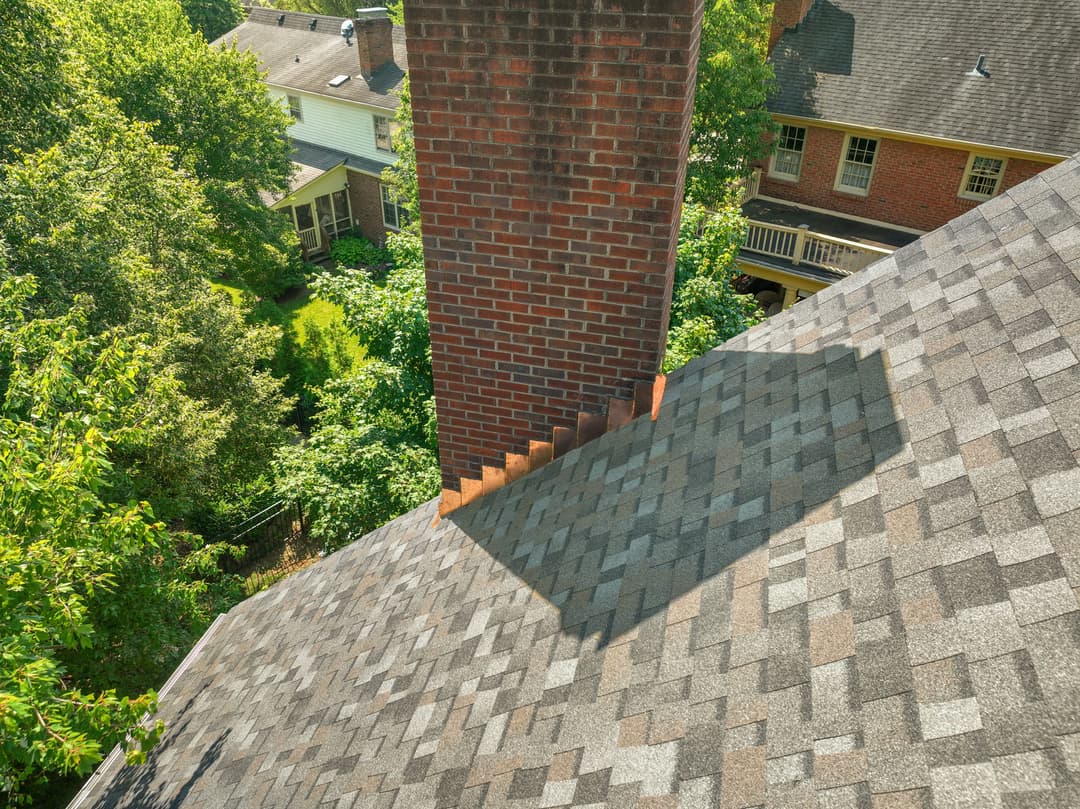 Aerial view of a chimney on a shingled roof surrounded by trees and houses.
