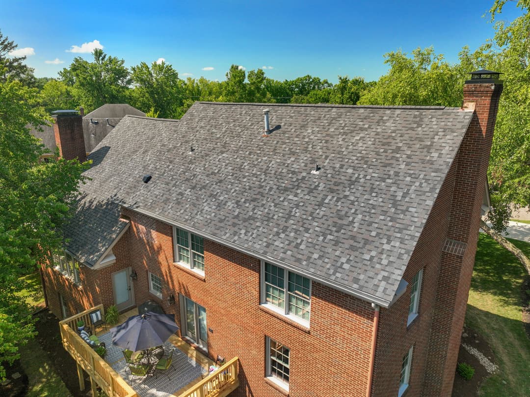 Two-story brick house with a new gray shingle roof and outdoor patio area surrounded by trees.