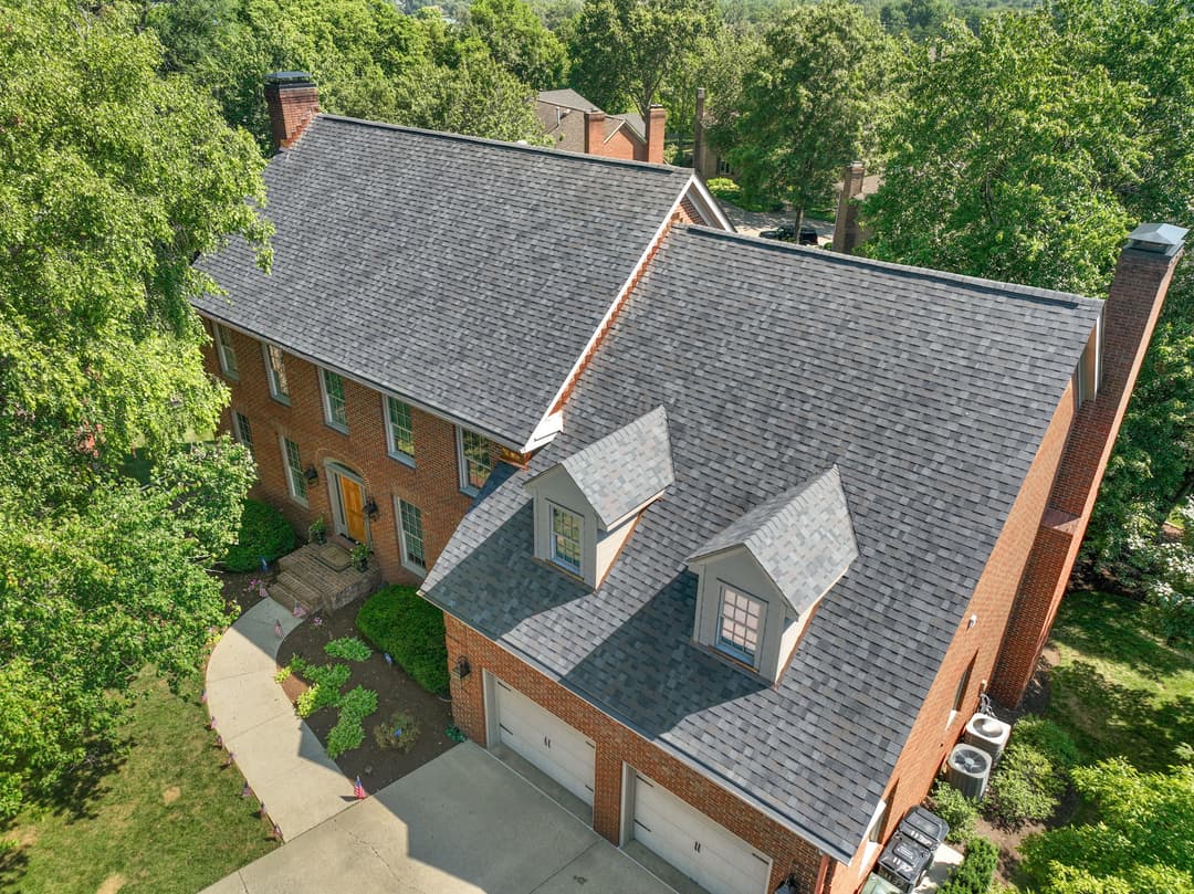 Aerial view of a brick house with gray shingles and landscaped yard, surrounded by trees.