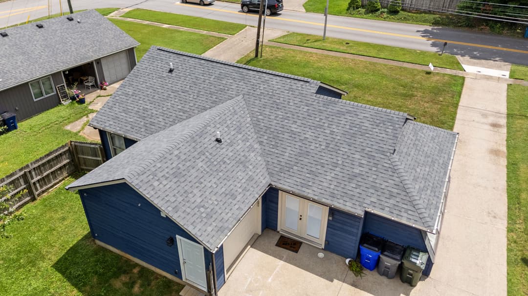 Aerial view of a blue house with a gray roof and driveway, surrounded by green grass.