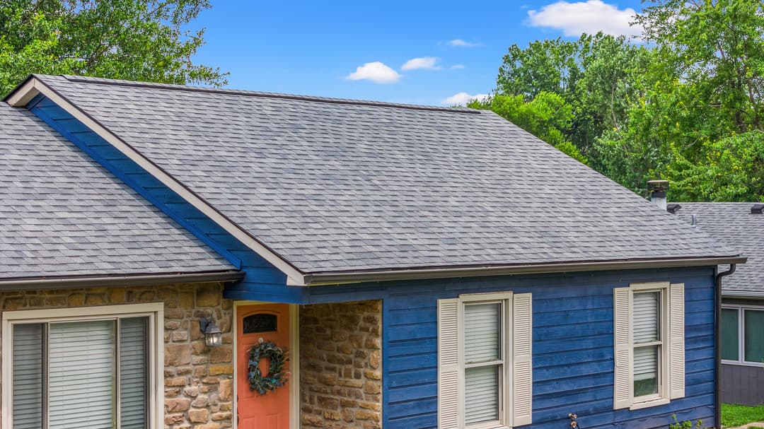 Blue house with stone accents and a modern roof, surrounded by lush greenery and clear skies.