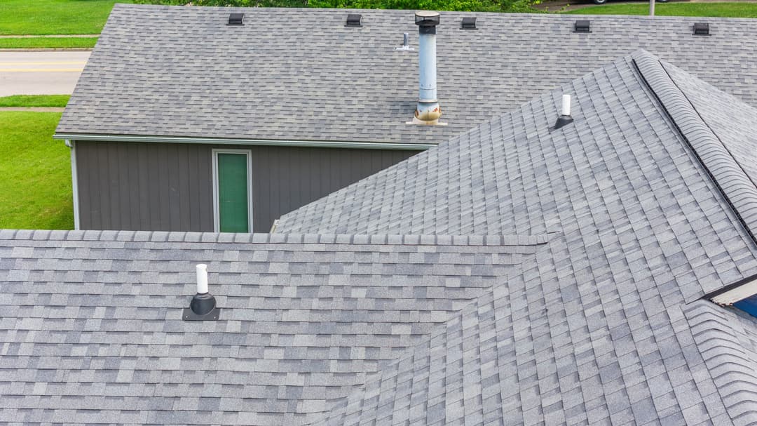 Aerial view of gray shingle roofs with ventilation pipes and green lawn background.