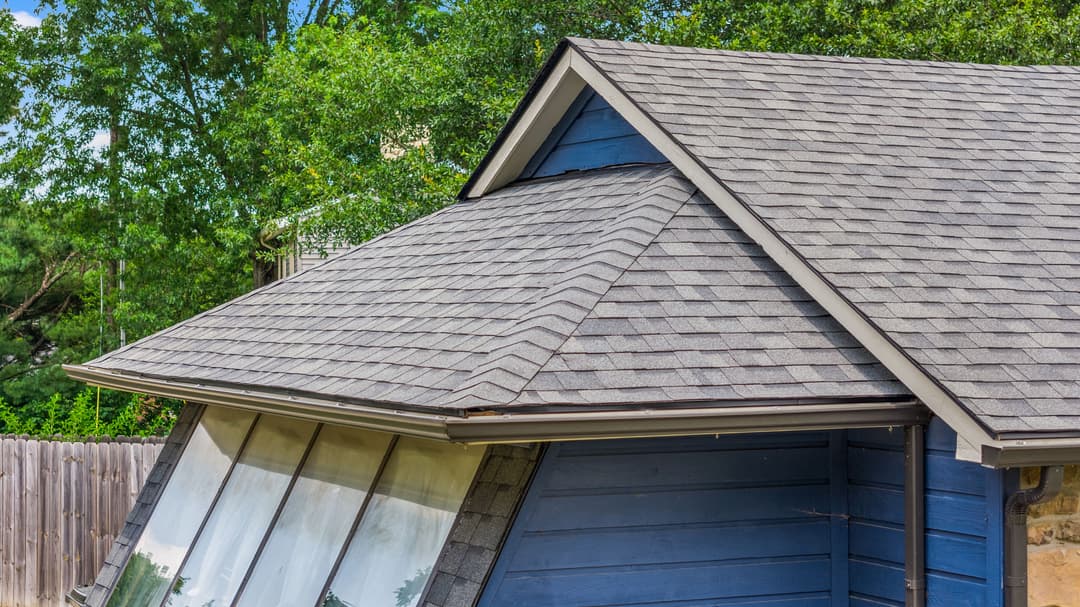 Roofline of a blue house with new shingles and greenery in the background.