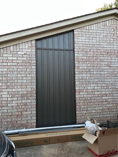 Exterior view of a modern vertical panel door on a brick wall, with nearby construction materials.